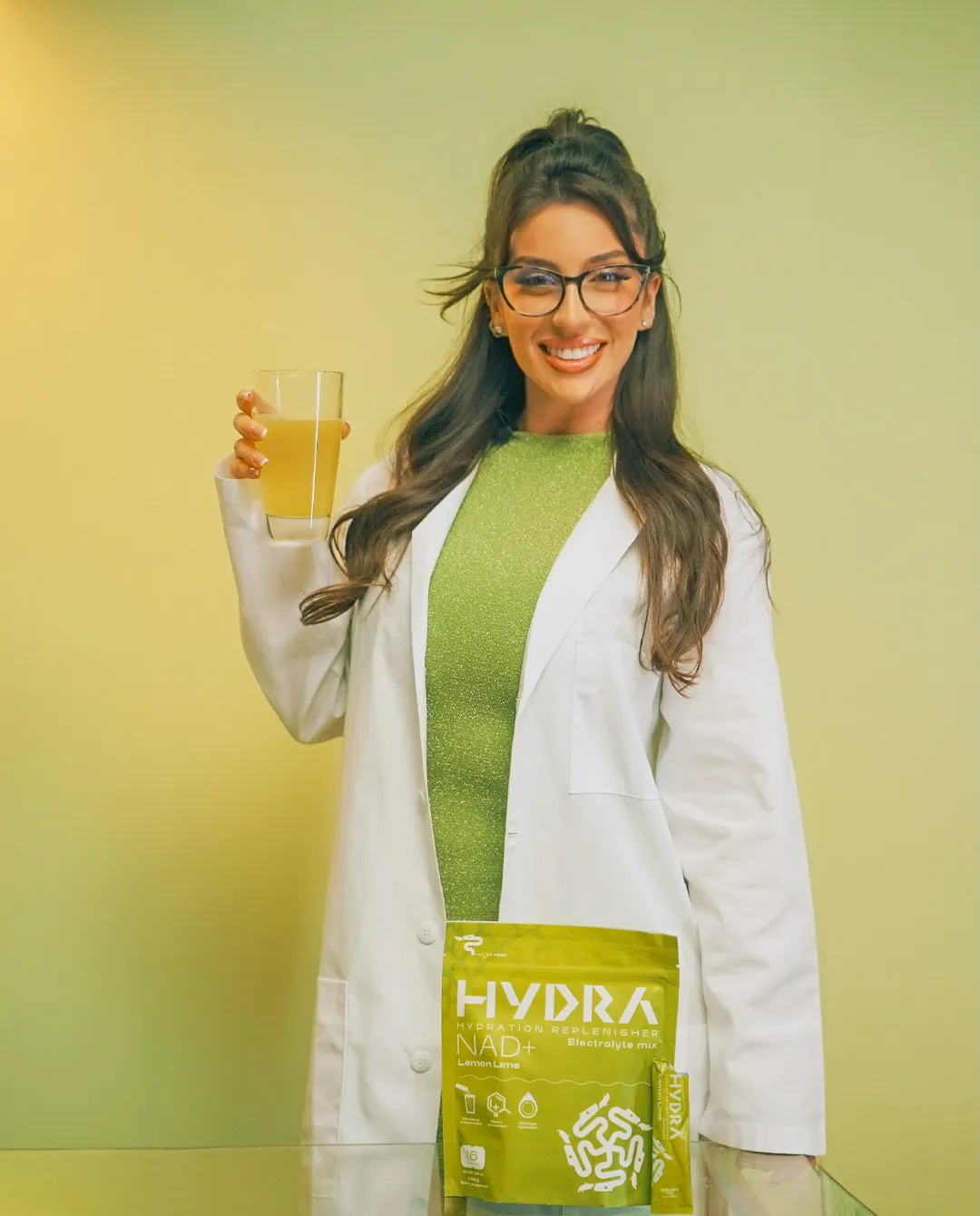 Lauren in a lab coat holding a glass and a pouch of Hydra Powders NAD+ electrolyte mix.