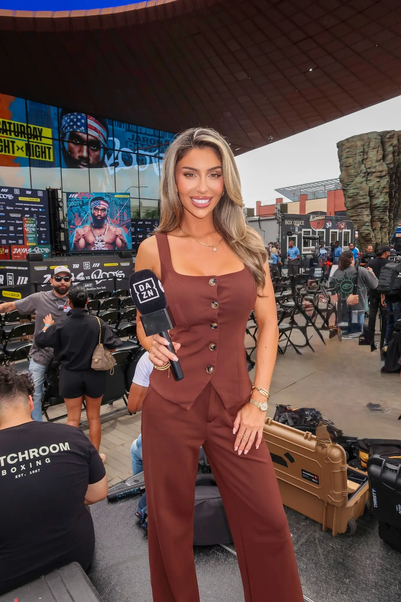 Lauren holding a DAZN microphone at the Zayas vs Ennis press event, with the fight artwork on screens behind her.